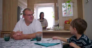 Anne, baba ve oğul birlikte otururken yemek servisi yapıyor. Sıcak bir mutfak ortamında gerçek bir aile etkileşimi.