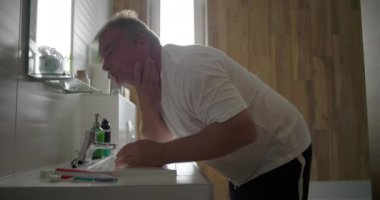 Yüzü akan suyun altında durulanan adam, lavaboya doğru eğilen, su sıçratmaya hazır eller, ahşap elementlerle dolu banyo ortamı, sabah bakım ritüeli.