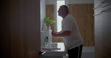 Adam banyoda tıraş oluyor, başını arkaya eğip çene altını tıraş ediyor, köpük uyguluyor, doğal banyo unsurları görülüyor, hijyen ve kişisel bakım üzerine odaklanıyor.