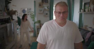 Evde hararetli tartışmalar yaşayan bir çift, erkek alaycı bir gülümseme sergilerken, kadın arka planda hayal kırıklığı içinde, evde gerginlik ve duygusal kopukluk sergiliyor.