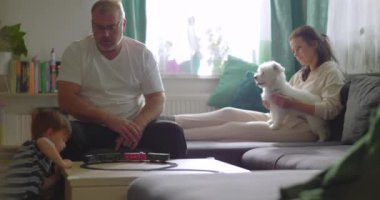 Baba sehpada genç bir çocukla oyuncak tren oyunu oynuyor, anne kanepede küçük beyaz bir köpekle gülümsüyor, neşeli bir aile, sıcak bir evde oturma odasında kaynaşıyor.
