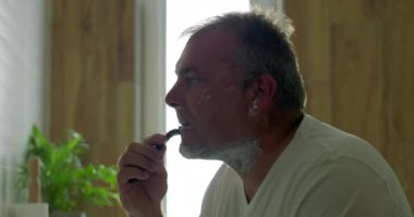 Banyoda tıraş olan bir adam, çene hizasında jilet kayması, tıraş köpüğü sürmesi, yan profil görünür, ayna yansıması, sabah bakım ritüelinin bir parçası.