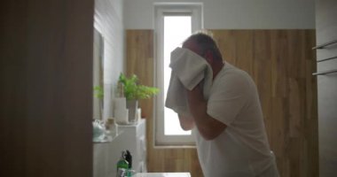 Beyaz havluyla yüzünü kurutan adam, duruladıktan sonra banyo lavabosunda duran, sabah bakım rutini, doğal ışık ve arka planda görünen ahşap banyo detayları.