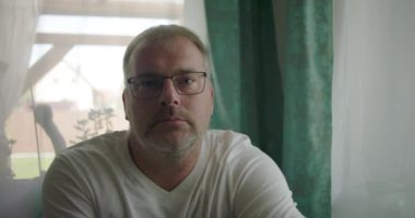 Pencerenin yanında oturan bir adam, sakin bir ev ortamında yumuşak perdelerle çevrili nötr ve içe dönük bir ifadeyle doğrudan kameraya bakıyor.