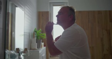 Adam yakın plan tıraş oluyor, boynun altından ustura süzülüyor, temiz tıraşa odaklanıyor, banyo arka planında doğal ahşap detaylar, hassas tımar anı, sakin bir atmosfer.