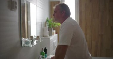 Banyo aynasında kendine bakan adam, düşünceli bir ifadeyle, ahşap ve beyaz fayanslarla dolu banyo içi, sessiz ve kişisel bir iç gözlem anı.