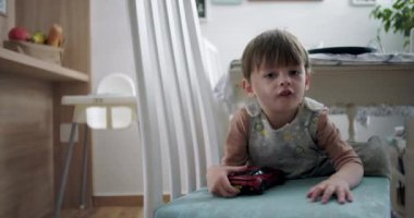 Çocuk sandalyeye yaslanıyor, kırmızı oyuncak bir araba tutuyor, meraklı bir bakışla, aile dostu bir ev ortamında yemek masasının altında.