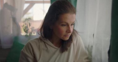 Bir pencerenin yanında oturan, düşünceli bir ifadeyle aşağı bakan bir kadın, yumuşak perdeler ve sakin bir evde yeşillik ile çevrili