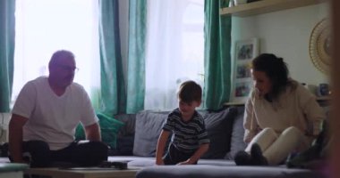 Aile rahat bir anı paylaşıyor, baba masaya oturuyor, genç çocuk öne eğiliyor, ve anne rahat oturma odasında kanepeye odaklanıyor.