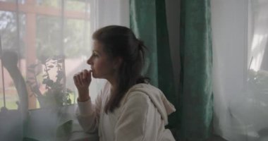 Pencere kenarında oturan bir kadın, düşünceli bir ifadeyle dışarı bakıyor, çevresi bitkiler ve yeşil perdelerle çevrili,