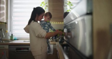 Anne bebeğini mutfakta tutarken çocuk sıcak bir yuva ortamında günlük aile rutini sırasında tezgaha, bağ kurma ve ebeveynlik anına doğru uzanır.