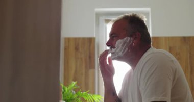 Yüze tıraş kremi süren, sabah tıraşına hazırlanan, temiz ve minimalist tuvaletlerde kendine bakım ve bakım rutinini vurgulayan bir adam.