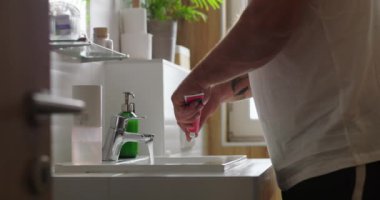 Banyodaki lavaboda diş fırçasına diş macunu süren bir adam, minimalist ve düzenli alandaki musluktan akan su, kişisel hijyen ve kendine bakımı vurgulayan bir adam.