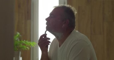 Usturayla tıraş olan, çene altını tıraş etmek için çenesini kaldıran, hassasiyete ve temiz bir sona odaklanan, doğal ahşap detaylarla dolu banyo ayarları ve ayna yansıması olan bir adam.