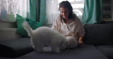 Kanepede oturan bir kadın rahat ev ortamında küçük beyaz bir köpeğe sarılıyor. Parlak pencere ve yastıklar içerideki sıcaklığı ve arkadaşlığı arttırıyor.