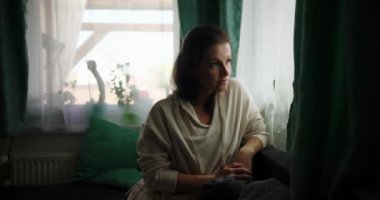 Kanepede oturan bir kadın, elleri kucağına yapışık, penceresinden dışarı bakan düşünceli bir yüz ifadesiyle, rahat bir ev içi ve yumuşak bir dekorla çevrili.