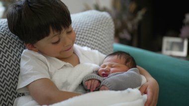 Küçük çocuk gülümserken yeni doğan kardeş, aile bağı, konforlu yuva kurma, kardeş sevgisi, hassas an, kapalı mekan sahnesi, aile bakımı, sevgi dolu bağ, yeni ekleme