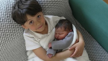 Neşeli bir yüzü olan genç bir çocuk yeni doğan kardeşini sıcak bir battaniyeye sarıp, saf mutluluk ve kardeşçe sevgi dolu bir an sergiliyor.