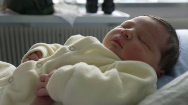 Yeni doğmuş bebek anne kollarında huzur içinde dinleniyor, rahat bir krem kıyafet giymiş. Yakın çekim sakin bir ifade ve sevgi dolu bir kucaklama, bebekle ilk zamanların sıcaklığını ve sakinliğini vurguluyor.