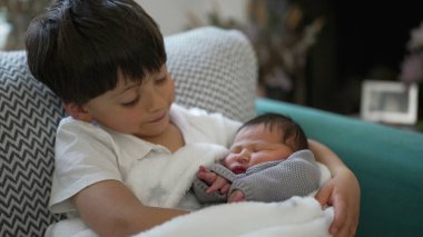Küçük çocuk gülümserken yeni doğan kardeş, aile bağı, konforlu yuva kurma, kardeş sevgisi, hassas an, kapalı mekan sahnesi, aile bakımı, sevgi dolu bağ, yeni ekleme