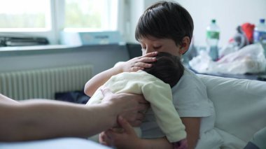 Çocuk, hastane odasında ebeveyn desteğiyle yeni doğan bebeği beşikte, kardeş bağını, aile sevgisini ve yetiştirme ortamını sembolize ediyor.