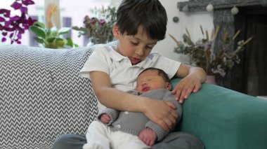 Genç bir çocuk, yeni doğmuş kardeşini sıcak bir ev dekoruyla çevrili bir kanepede sevgiyle kucaklıyor, aile sevgisini ve yakınlığını ön plana çıkarıyor. Kardeş sevgisi, ilgi ve aile birliği temalarını vurgular