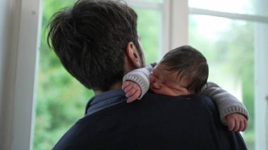 Yeni doğmuş bebek babasının omzunda dinleniyor, rahat örülmüş bir kazak giyiyor. Baba pencereye doğru döner, aralarındaki rahatlık ve güven dolu kucaklaşmayı vurgular.