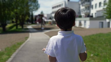 Bir yolun üzerinde duran, çocuk parkına ve konut binalarına bakan, güvenli, banliyö ortamında merak ve keşifleri ön plana çıkaran genç bir çocuğun arka görüntüsü