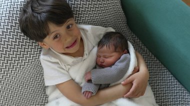 Neşeli bir yüzü olan genç bir çocuk yeni doğan kardeşini sıcak bir battaniyeye sarıp, saf mutluluk ve kardeşçe sevgi dolu bir an sergiliyor.