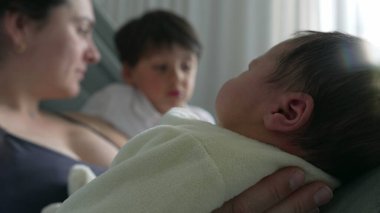 Anne ve büyük kardeş yeni doğan bebeğe bakıyor, aile içi sevgi ve hastane yatağında bir bağ yakalıyorlar. Anne, büyük çocuk ve yeni doğan arasındaki hassas etkileşim ve bağ