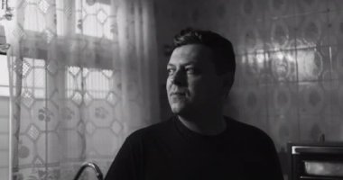 Mutfakta duran, pencereden dışarı bakan, siyah-beyaz ayar, içsel ve yansıtıcı ruh hali, sakin ev ortamında klasik detaylar.