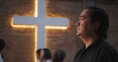 Kilisenin içinde derin bir tefekkür ve dua içinde yukarı bakan bir adam, arka plandaki tuğla duvarda parlayan bir haç, maneviyat ve inancı çağrıştırıyor.