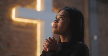 Genç bir kadın dua ederken elini sıkar ve kilisenin içine doğru bakar, arka plandaki tuğla duvarda parlayan bir haçla, inanç ve ruhaniliği çağrıştırır.