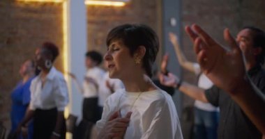 Kapalı gözlerle dua eden ve çeşitli grup ibadet ortamlarında el kaldıran kadın, arka plan, inanç, maneviyat ve toplumsal adanmışlık alanlarında aydınlatılmış haç.