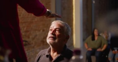 Kilisenin kutsaması sırasında diz çökmüş yaşlı bir adam, ruhani lider tarafından alnına konmuş bir el, inanç, bağlılık ve tevazu ifadesi, kapalı mekanda dini bir toplantı.