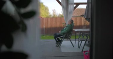 Arka bahçede terasta oturan, kolları bağlı bir sandalyede dinlenen sakin bir bahçe ortamında sakin bir anın tadını çıkaran bir adam.