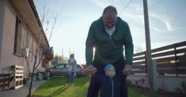 Anne uzaktan izlerken, baba bebeğin çimenlerde yürümesine yardım ediyor. Aile birlikte olmanın ve doğal ortamlarda öğrenmenin tadını çıkarıyor.