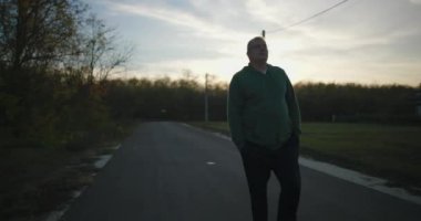 Gün batımında boş bir kırsal yolda yürüyen bir adam, elleri ceplerinde, huzurlu bir kırsal bölgede sonbahar ağaçları ve açık gökyüzü ile huzurlu bir akşamın tadını çıkarıyor.