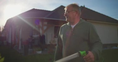 Bahçe hortumunu arka bahçeyi sulamak için kullanan bir adam varoş ortamında çimlere doğru kıvrılan su akıntısı arka planda görünen ev ve veranda, sakin bir an.