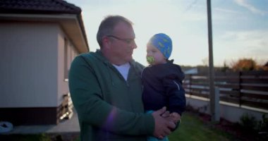 Baba bebeği nazikçe kucağında tutuyor, evin arka bahçesinde duruyor, dışarıda sakin ve sıcak ebeveyn anlarının tadını çıkarıyor.