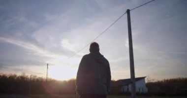 Kırsal bir yolda yürüyen bir adam, uzaklara bakıyor, gün batımında ufka bakıyor, sakin ve sakin bir atmosfer, uzaktaki evleri ve ağaçları olan bir kırsal alan.