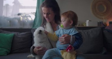 Anne koltukta tüylü beyaz bir köpeği rahatlatırken bebeğini kucağında tutuyor. Bebek emzik ve sıcak bir aile ortamında samimi bir aile anı yaşıyor.