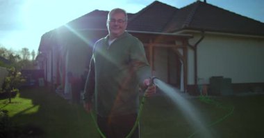Bahçe hortumuyla çimenleri sularken gülümseyen bir adam, banliyö evlerinde rahat bir atmosfer, arka planda ev, veranda ve yeşillik olan bir ortam.