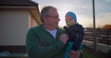 Baba, bebeğini emzik ile arka bahçede tutuyor. Çocuk kıyafetlerini nazikçe ayarlıyor. Huzurlu bir aile ortamında sevgi ve sevgi gösterisi yapıyor.