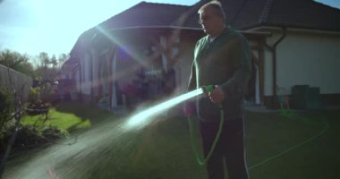 Bahçede bahçe hortumuyla çimenleri sulayan bir adam, su püskürtme ile sıradan bir an, banliyö ortamında konut ve yeşillik, rahat ve sakin bir atmosfer.