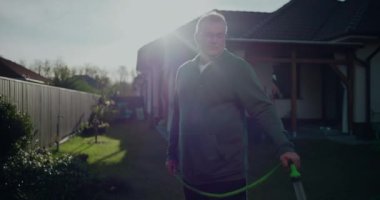 Bahçe hortumunu arka bahçede tutan adam, konut evinin yanında duran, yeşil, veranda ve çimenli, rahat açık hava aktivitesi olan banliyö ortamında sıradan bir an.