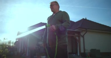 Arka bahçeye bahçe hortumuyla su fışkırtan bir adam, arka planda konut ve yeşillik, rahat banliyö ortamında günlük aktiviteler ve parlak gün ışığı.