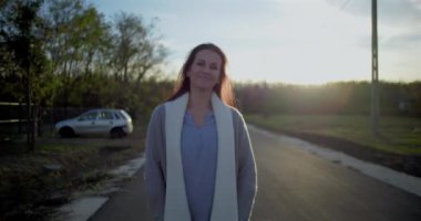 Sessiz, kırsal bir yolda yürüyen bir kadın, yüzü öne dönük, arka planda sakin bir gün batımı, park halindeki arabasıyla kırsal bir ortam, sakin bir ortam.