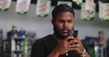 Afrika kökenli genç adam dikkatli bir şekilde bir mağaza ortamında akıllı telefon ekranına odaklanmış, dijital bağlılık ve teknolojiye modern bağımlılığı göstermektedir