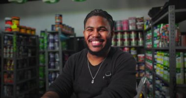 Boya dükkanında çalışan gülümseyen adam, arka planda organize boya kutularıyla tezgahta oturan profesyonel ve cana yakın bir çalışan.